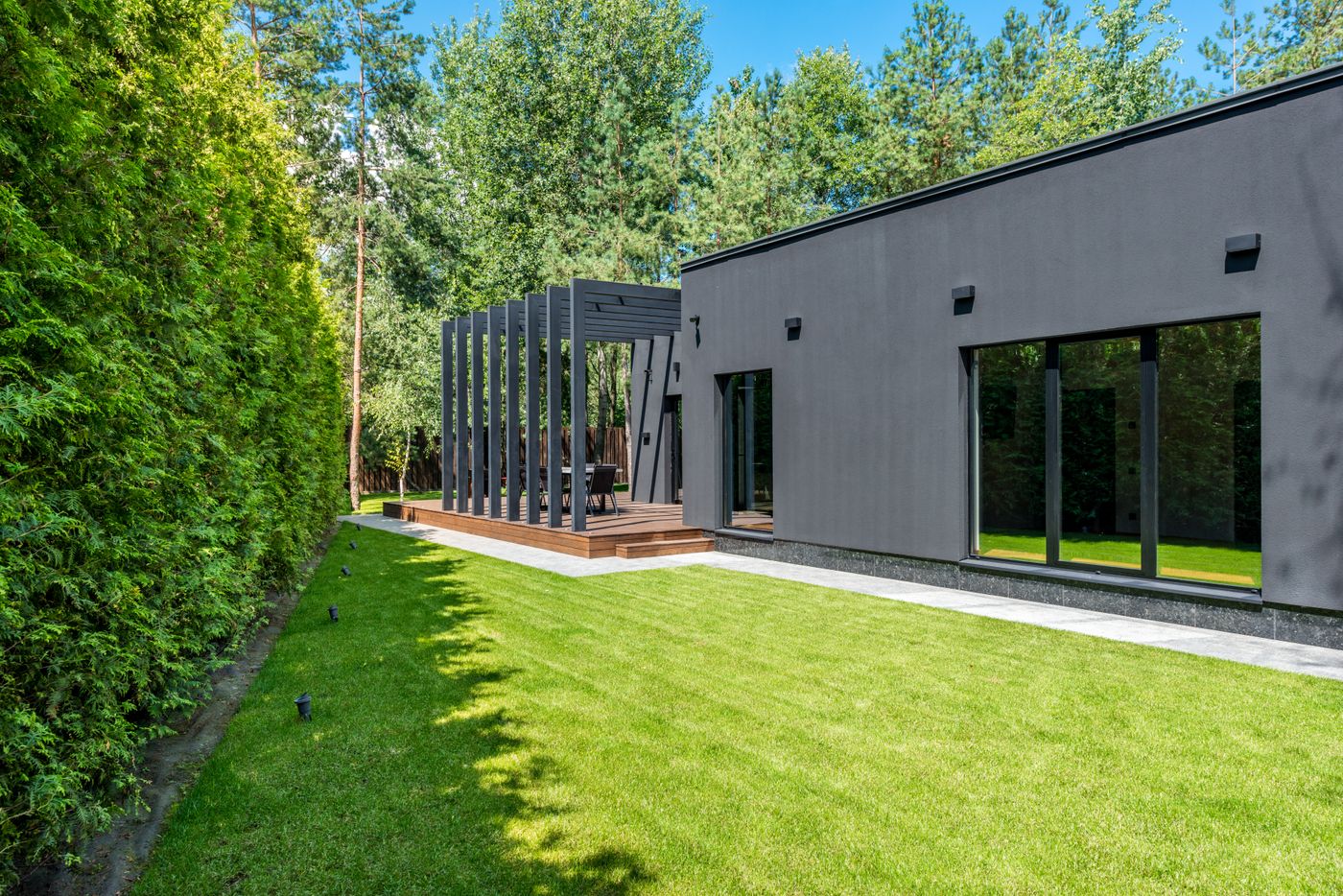 Modern gray house with wooden deck and pergola surrounded by green lawn and forest trees