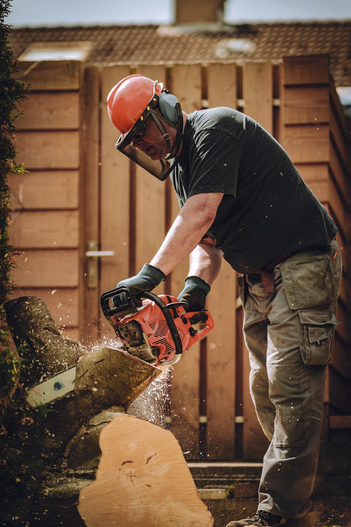 Worker in red helmet operating chainsaw to cut log with brick building in background